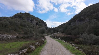 Buracas do Casmilo, Condeixa, Portekiz yakınlarındaki kayalık yamaçlı dar bir vadiden geçen toprak yol. Zeytin ağaçları ve dağınık bulutlu mavi gökyüzünün altında dağılmış taş duvarlar..
