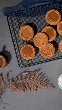 Caramel tarts on a cooling rack with a jar of caramel sauce, a bowl of salt, and a dried fern leaf on a dark surface, creating a rustic aesthetic clipart