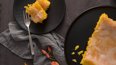 Lemon cake slice on black plate with fork, grey cloth, and flowers; another plate with larger cake portion; dark background; crumbs scattered clipart