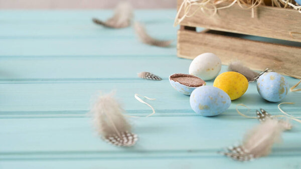 Easter eggs and feathers on a light blue wooden surface, with a wooden crate filled with straw in the background, creating a festive and rustic scene