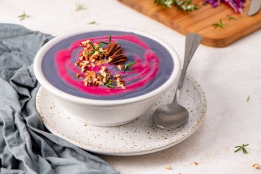 Purple soup with pink swirl, topped with pecans and rosemary, in a white speckled bowl on a matching plate, with a spoon and gray cloth nearby clipart