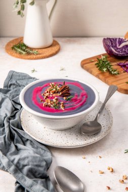 Purple soup garnished with pecans and rosemary in a white bowl on a speckled plate, accompanied by a spoon and a gray napkin, with a cutting board and herbs in the background clipart