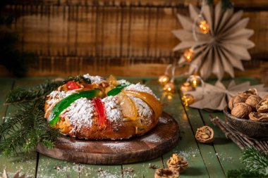 Festive holiday bread topped with candied fruits, powdered sugar, and almonds, set on a rustic wooden table surrounded by Christmas decor. clipart