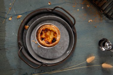 Tek bir Pastel de Nata, kırsal bir metal tepside dinleniyor, üzerine tarçın serpiştirilmiş..