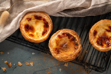 Altın pastel de nata hamur işleri, siyah bir tel askıda soğuyor, yanında, kırsal bir yüzey üzerindeki kırıntılarla çevrili, dokulu keten bir kumaş var..