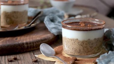 Üç lezzetli tiramisu, şeffaf cam kaplarda, kremalı yulaf, kahve ve kakao tozuyla kaplı, üzerinde çikolata parçaları ve kahve çekirdekleri olan. Mavi bir bez ve kaşıkla ahşap bir tepsiye konmuş, harika bir ikram için mükemmel..