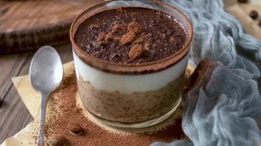 Bir gecelik tiramisu servisi şeffaf cam bir kapta, kremalı yulaf, kahve ve kakao tozuyla kaplı, üzerinde çikolata parçaları ve kahve çekirdekleri olan..