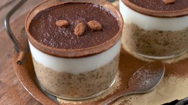 Kremalı yulaf, kahve ve kakao tozu katmanlı, üzerinde çikolata parçaları ve kahve çekirdekleri olan, temiz cam kaplarda iki lezzetli tiramisu. Mavi bir bez ve kaşıkla ahşap bir tepsiye konmuş, harika bir ikram için mükemmel..