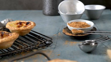 Soğutma rafında iki Portekizli muhallebi tartı, tarçın kaplı kaşıklar ve dokulu mavi bir yüzey üzerinde pişirme konserveleri.