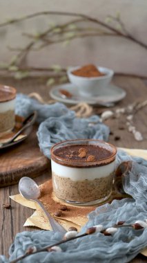 Lezzetli bir tiramisu, üzerinde çikolata parçaları ve kahve çekirdekleri olan kremalı yulaf, kahve ve kakao tozu katmanlı cam bir kapta bir gecelik yulaf. Mavi bir bez ve kaşıkla ahşap bir masaya kurulmuş, harika bir ziyafet için mükemmel..