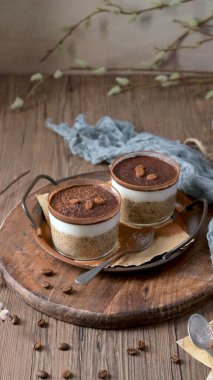 Kremalı yulaf, kahve ve kakao tozuyla kaplı cam kaplarda iki lezzetli tiramisu. Çikolata parçacıkları ve kahve çekirdekleri ile süslenmiş, bunlar ahşap tepside mavi bir bez ve kaşık ile hazırlanmış, zevk için mükemmel.