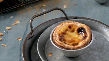 Tek bir Portekiz kremalı turtası metal bir teneke kutunun içinde yuvarlak metal bir tepside oturur ve etrafına kırıntılar saçılmıştır.