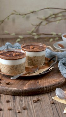 Kremalı yulaf, kahve ve kakao tozu katmanlı, üzerinde çikolata parçaları ve kahve çekirdekleri olan, temiz cam kaplarda iki lezzetli tiramisu. Mavi bir bez ve kaşıkla ahşap bir tepsiye konmuş, harika bir ikram için mükemmel..