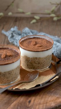 Kremalı yulaf, kahve ve kakao tozu katmanlı, üzerinde çikolata parçaları ve kahve çekirdekleri olan, temiz cam kaplarda iki lezzetli tiramisu. Mavi bir bez ve kaşıkla ahşap bir tepsiye konmuş, harika bir ikram için mükemmel..