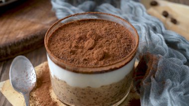 A single serving of tiramisu overnight oats in a clear glass container, layered with creamy oats, coffee, and cocoa powder, topped with chocolate pieces and coffee beans. Set on a wooden table with a blue cloth and a spoon, perfect for a delightful t
