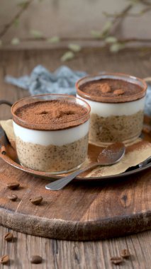 Kremalı yulaf, kahve ve kakao tozu katmanlı, üzerinde çikolata parçaları ve kahve çekirdekleri olan, temiz cam kaplarda iki lezzetli tiramisu. Mavi bir bez ve kaşıkla ahşap bir tepsiye konmuş, harika bir ikram için mükemmel..