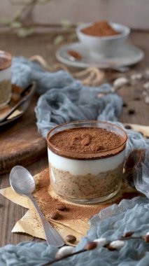 Sütlü yulaf, kahve ve çikolata parçacıkları ve kahve çekirdekleri ile kaplı şeffaf cam bir kapta nefis bir tiramisu. Mavi bir bez ve kaşıkla ahşap bir masaya kurulmuş, harika bir ziyafet için mükemmel..