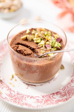 Dilimlenmiş fıstıkla kaplı tek bir porsiyon lezzetli çikolatalı tatlı beyaz ve kırmızı tabakta bir kaşıkla berrak bir bardakta sunuluyor.