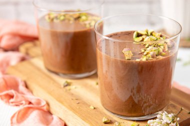 İki bardak çikolatalı avokado mousse, ahşap bir tahtanın üzerinde pembe peçeteyle süslenmiş fıstıkla süslenmiş.