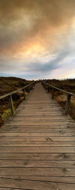 Tahta bir sahil yolu, öğleden sonra güneşinin sıcak renkleriyle dramatik ve dumanlı bir gökyüzünün altında uzanıyor..