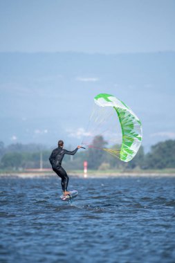 Dalgıç kıyafetli orta yaşlı bir adam, yeşil ve beyaz bir uçurtmayla birlikte, uzaktaki ağaçlarla kaplı bir sahilde su folyosunun üzerinde süzülüyor.