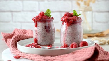 İki kavanoz chia tohumu pudingi parlak ahududu kompostosu ile kaplı ve taze nane yapraklarıyla süslenmiş beyaz bir tabakta gösteriliyor. Tatlı beyaz mermer bir yüzeyde sunuluyor. Pembe desenli bir peçete ve sırtında kırsal tuğla bir duvar var.