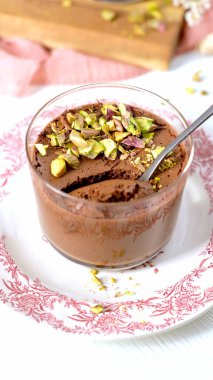 Süslü beyaz bir tabakta kırmızı çiçek desenli bir bardak zengin çikolatalı mus. Yumuşak, hafif tatlının içinde bir kaşık dinleniyor, yumuşak dokusunu sergiliyor ve üzerine bol miktarda ezilmiş şam fıstığı serpiştiriyor..