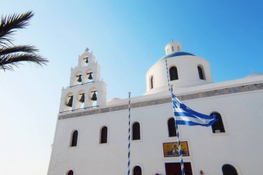 The Orthodox Church of Panagia Akathistos Hymn Holy with the Greek flag in front of the facade. Oia, Santorini. Santorini Island, Greece. clipart