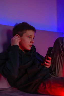 Teenage boy using phone and headphone in his room with neon light.