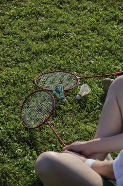 İki badminton raketi ve mekik horozu çimlerin üzerinde yatıyor. Arka planda bir kişi oturuyor..