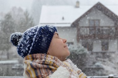 Neşeli bir birey dışarıda duruyor, ılık bir şapka ve renkli bir eşarp giyerken hafif kar yağışını kucaklıyor, sakin bir kış ayarıyla çevrili..