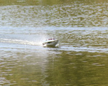 Durgun sularda siyah çizgili, beyaz, elektrikle çalışan, radyo kontrollü bir tekne, ardında hafif bir iz bırakarak hızla ilerliyor. Huzurlu sahnede bulutsuz mavi bir gökyüzü var..