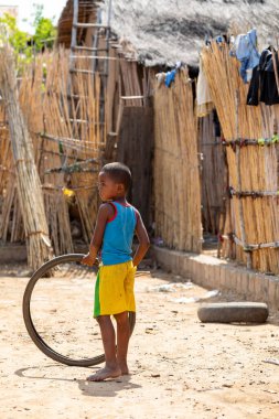 Miandrivazo, Madagaskar - 27 Kasım 2022: Genç bir çocuk toprak bir yolda dururken eski bir bisiklet lastiğiyle oynuyor.