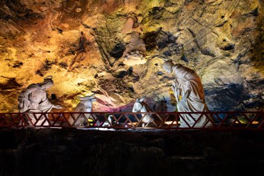 Nativity scene with stone sculptures of Mary, Joseph, baby Jesus in a manger, and an angel above, set against the illuminated rock wall of the Salt Cathedral of Zipaquira, Colombia. clipart