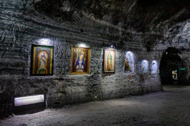 Religious artwork and statues of the Virgin Mary displayed on the textured stone walls of the Salt Cathedral of Zipaquira, Colombia, illuminated by soft lighting in a dimly lit underground chamber. clipart