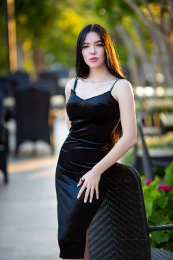 Beautiful sexy young long-haired brunette caucasian girl in slimming black dress leaning on a chair and attractively posing at Antalya hotel's terrace with softly blurred greenish background