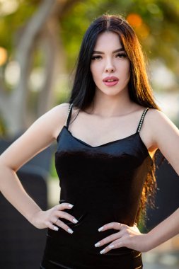 Beautiful sexy young long-haired brunette caucasian girl in slimming black dress attractively posing at Antalya hotel's terrace with softly blurred greenish background