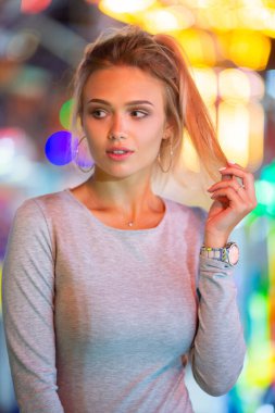 Portrait of a beautiful slim blonde caucasian girl looking left sexually posing, shot in amusement park with colored bulb lights at the background blurred in huge bokeh and lens flares