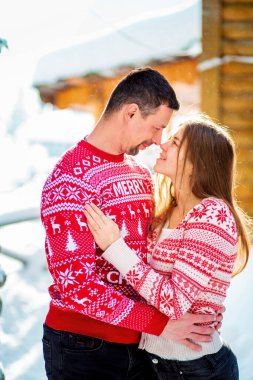 Engagement session, love story of a young straight couple hugging and kissing in Christmas red sweaters and jeans happily looking at each other in snowy landscape near a wooden house and pine trees