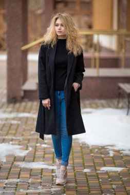 Siyah trençkotlu ve mavi kotlu güzel, genç, beyaz, kıvırcık, kıvırcık, sarışın bir kızın tam boy portresi.