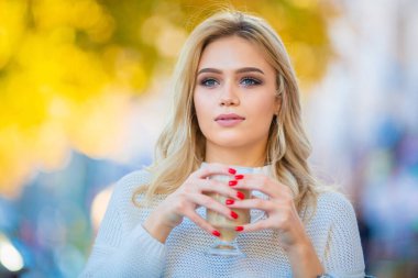 Elinde bir fincan kahve, büyük gümüş küpeler ve kırmızı manikür, bulanık mavi sonbahar arkaplanı, gülümseyen ve elinde bir fincan kahve tutan güzel, genç, beyaz bir kızın yatay portresi.