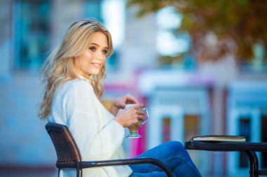 Kafkasyalı güzel bir kızın yatay portresi gülümseyen ve elinde bir fincan kahve, masanın üstünde kitap, büyük gümüş küpeler ve kırmızı manikür, bulanık mavi sonbahar arkaplanı