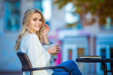 Kafkasyalı güzel bir kızın yatay portresi gülümseyen ve elinde bir fincan kahve, masanın üstünde kitap, büyük gümüş küpeler ve kırmızı manikür, bulanık mavi sonbahar arkaplanı