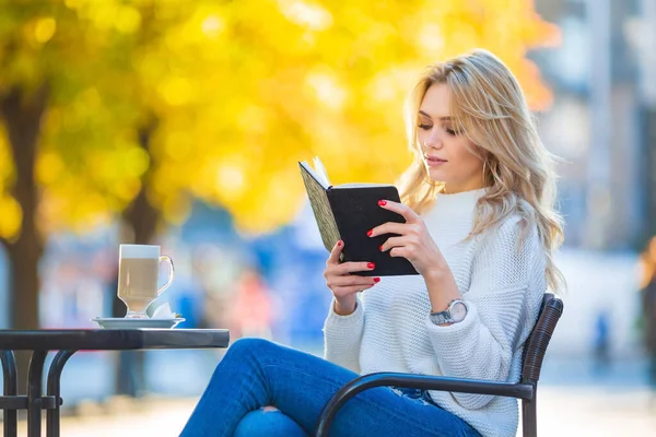一个漂亮的年轻的高加索金发姑娘拿着一本书看书 桌上放着一杯咖啡 大大的银耳环和红色的指甲 模糊的蓝黄相间的秋天背景的画像 — 图库照片