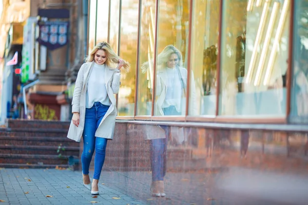 全长肖像一个美丽的年轻的高加索金发姑娘笑着走在商店橱窗附近整理她的头发 蓝色牛仔裤和米黄色的风衣 模糊的蓝色秋天的背景 — 图库照片