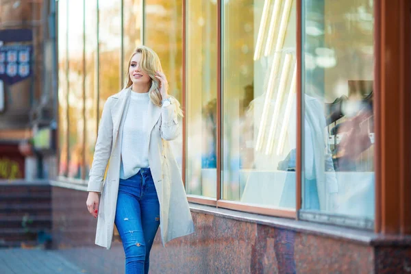 一个美丽的年轻的高加索金发姑娘笑着走在商店橱窗边整理她的头发 蓝色牛仔裤和米黄色的风衣 模糊的蓝色秋天背景的横向肖像 — 图库照片