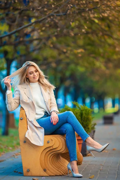 청바지와 베이지 코트를 벤치에 편안하게 아름답고 금발의 미소짓는 소녀의 푸르스름 — 스톡 사진