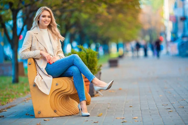 一个年轻美丽的高加索金发女郎坐在木制长椅上 穿着蓝色牛仔裤和米黄色的风衣 背景模糊的蓝色秋天的横向肖像 — 图库照片
