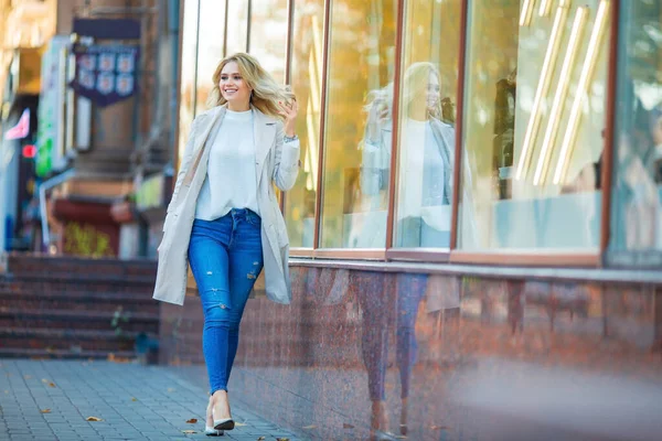 一个美丽的年轻的高加索金发姑娘笑着走在商店橱窗边整理她的头发 蓝色牛仔裤和米黄色的风衣 模糊的蓝色秋天背景的横向肖像 — 图库照片