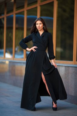 Uzun siyah tasarımcı elbisesi ve yüksek topuklu topuklu güzel, ince, beyaz, esmer bir kızın tam boy portresi. Hafif bulanık arka planlı, ritmik vitrinlerin yanında poz veriyor.